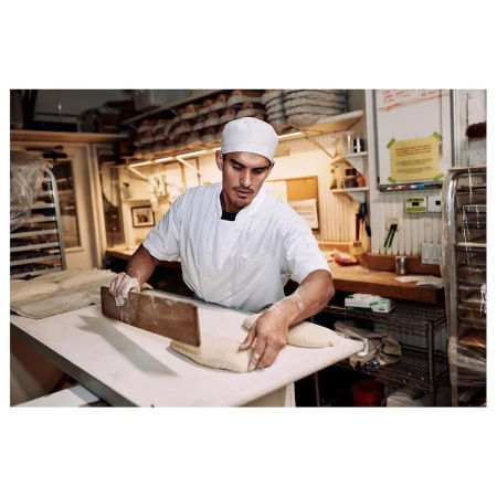 Adult male baker shaping dough at a bakery with white walls and stainless steel equipment; wearing a white baker's uniform and hat.