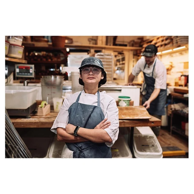 Woman in work apron and baseball cap poses in commercial kitchen, with a man working in background. Stainless steel surfaces.