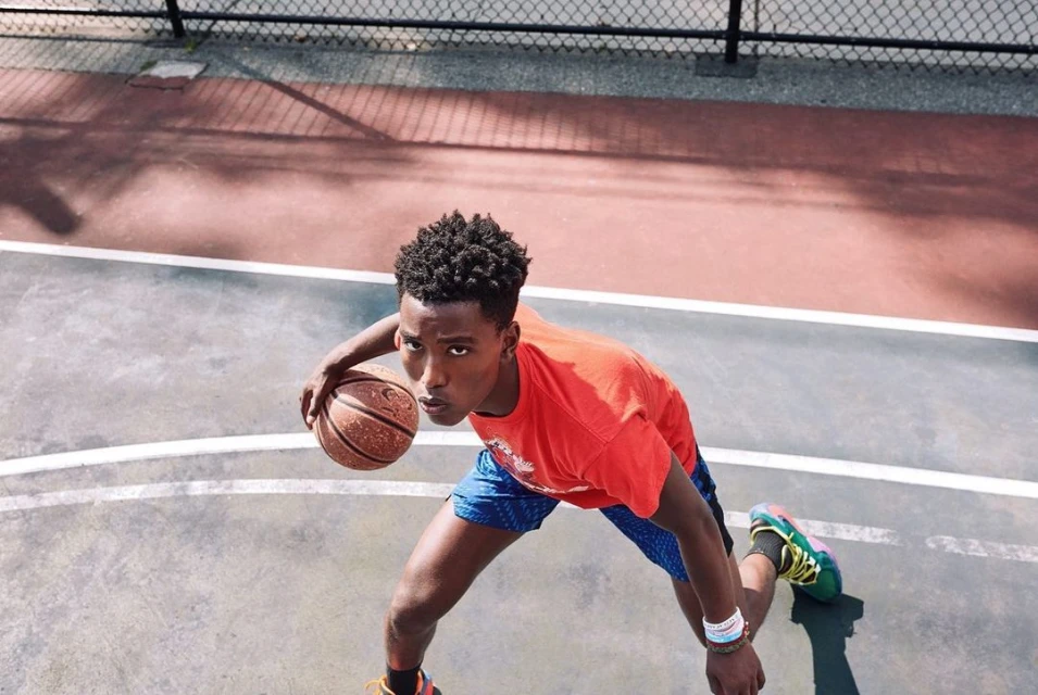 An African American man in an orange t-shirt dribbles a basketball on an outdoor court, shot from a high angle on a sunny day.