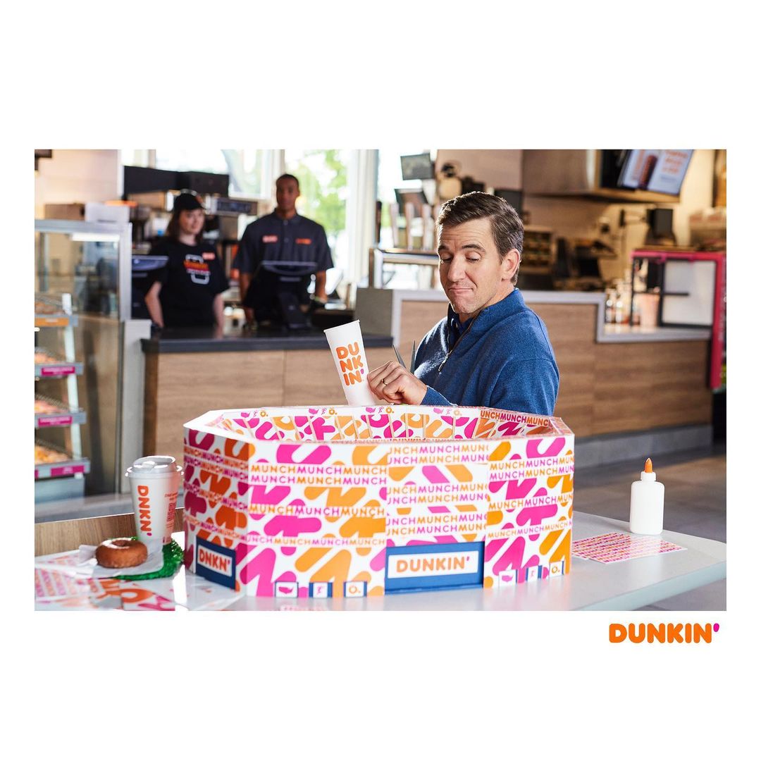 Adult male crafting a box labeled Dunkin' Donuts, two employees stand behind the counter inside the donut shop. 