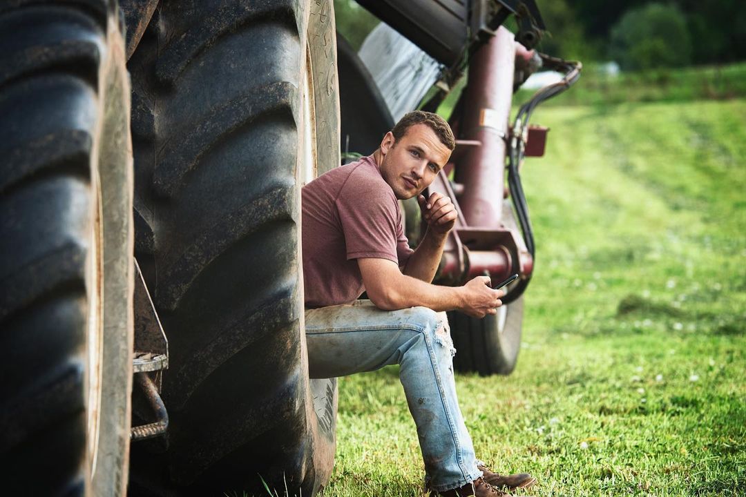 Man sits pensively by a tractor tire on a rural farm, gazing toward the camera while holding a cell phone in a grassy field.