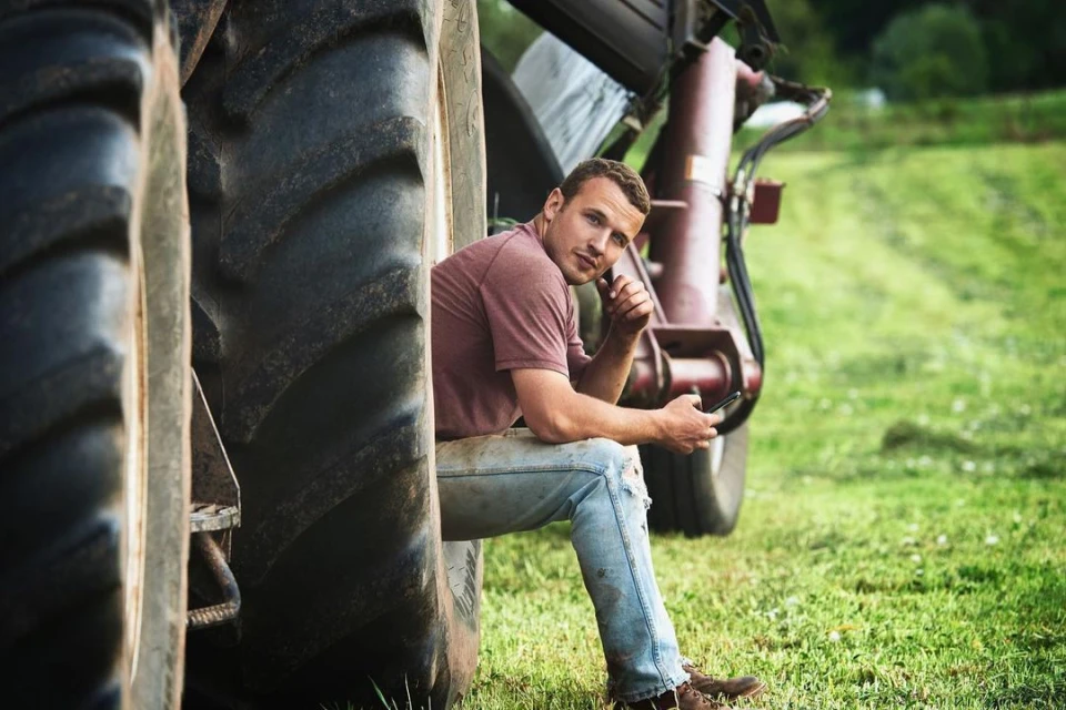 Man sits pensively by a tractor tire on a rural farm, gazing toward the camera while holding a cell phone in a grassy field.