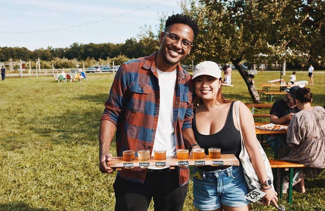 A smiling couple at an outdoor event holds a tasting flight of beers on a wooden paddle; other people, tables, and trees visible.