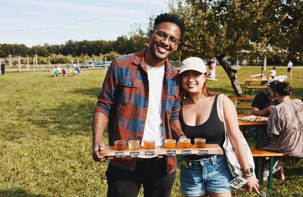 A smiling couple at an outdoor event holds a tasting flight of beers on a wooden paddle; other people, tables, and trees visible.