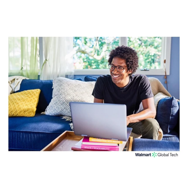 Smiling man with glasses works on a laptop on a blue couch with throw pillows, sitting near a stack of books and a window.