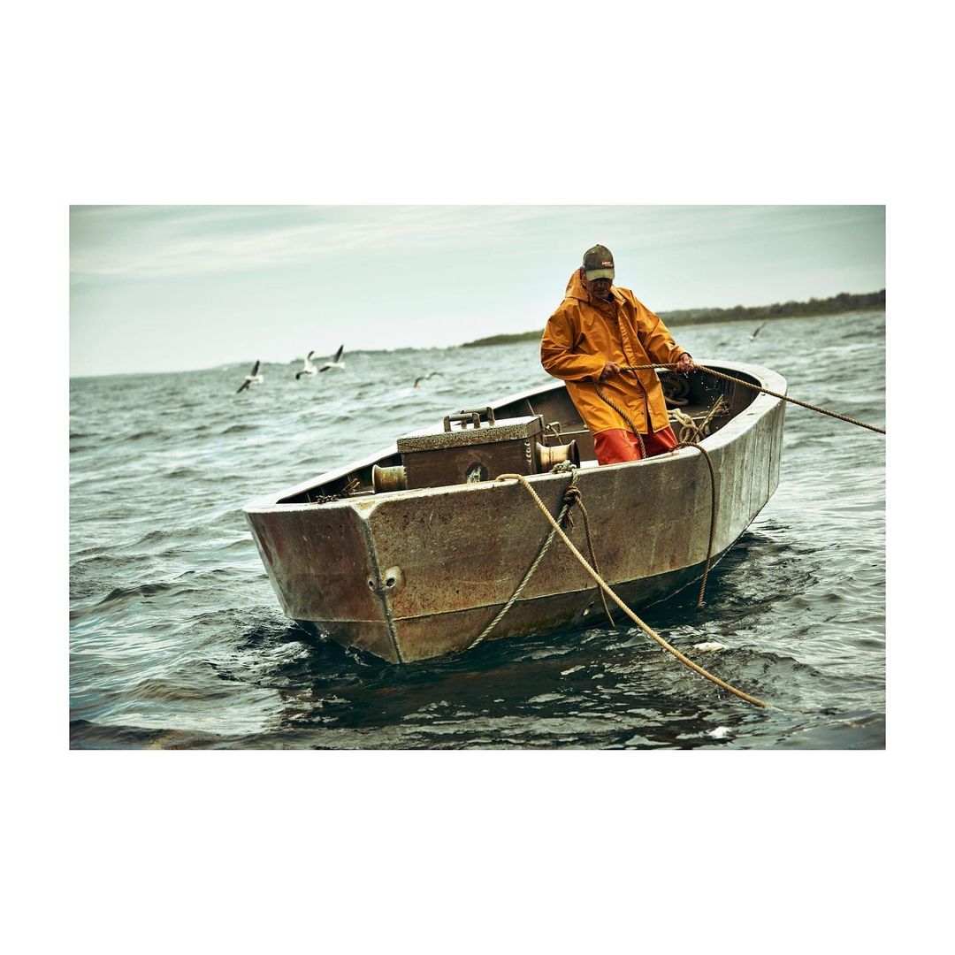 An adult male in an orange waterproof jacket rows an old, rusty boat on a choppy sea beneath an overcast sky with distant birds.