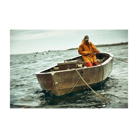 An adult male in an orange waterproof jacket rows an old, rusty boat on a choppy sea beneath an overcast sky with distant birds.