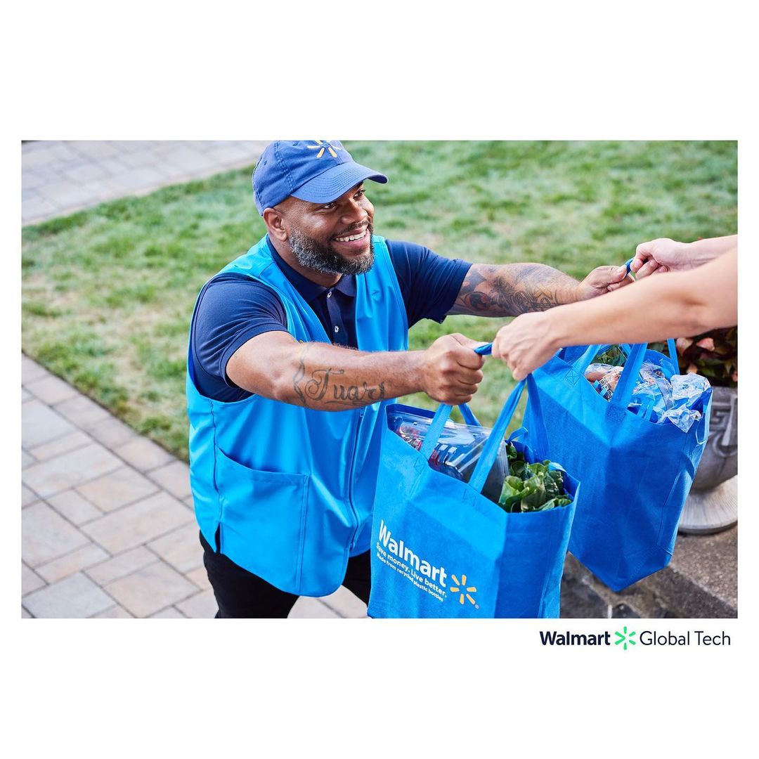 Friendly delivery person in blue uniform hands blue reusable grocery bags to a customer outside, creating a positive and convenient shopping experience.