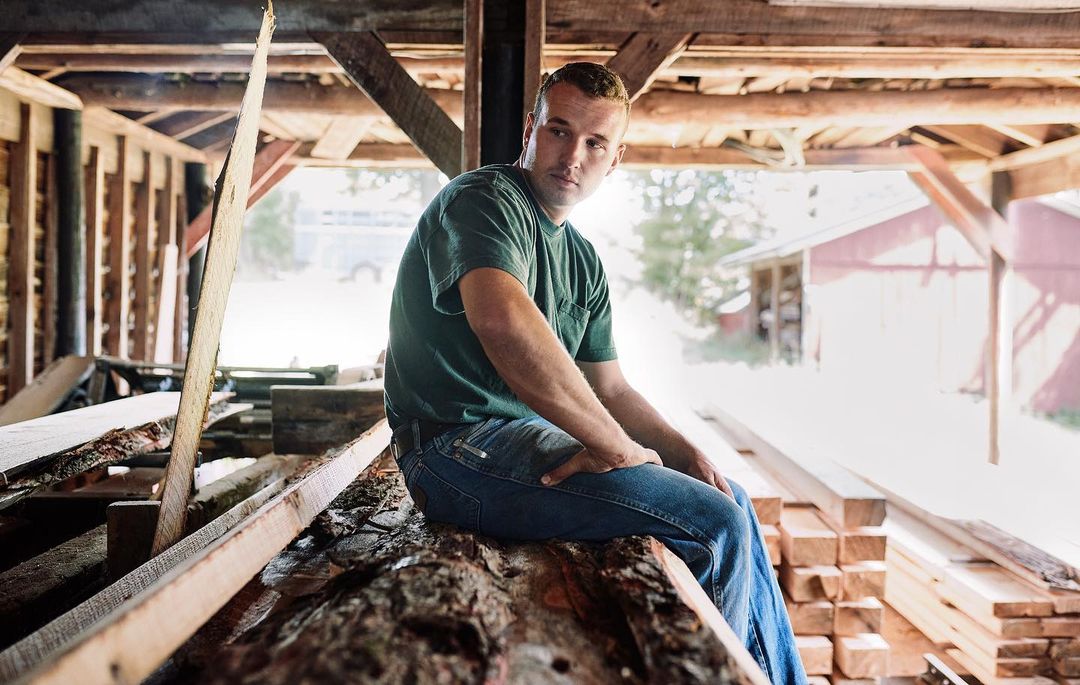 Adult male sits on a stack of logs inside a wooden structure, wearing a green tee shirt and jeans with a building in the background.