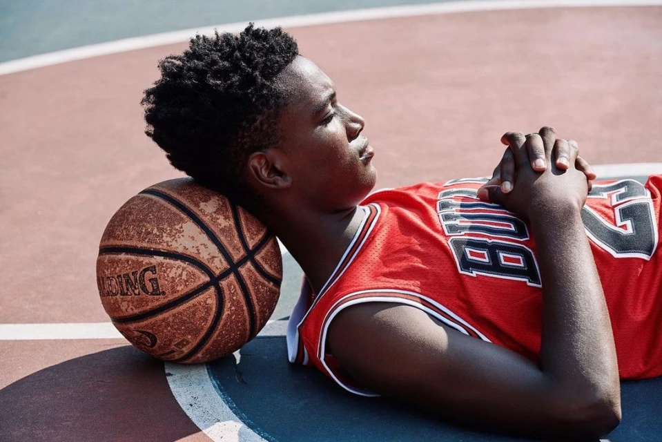 Teenage African-American basketball player relaxes on the court, resting his head on his ball while wearing a red jersey.