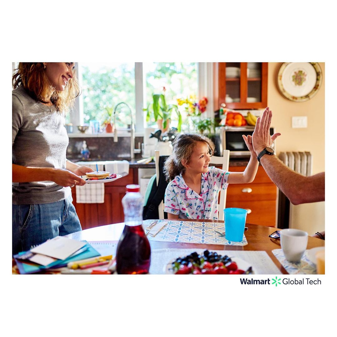 A family interacts at a kitchen table with breakfast items including fruit, pancakes, and syrup, with natural light from a nearby window.