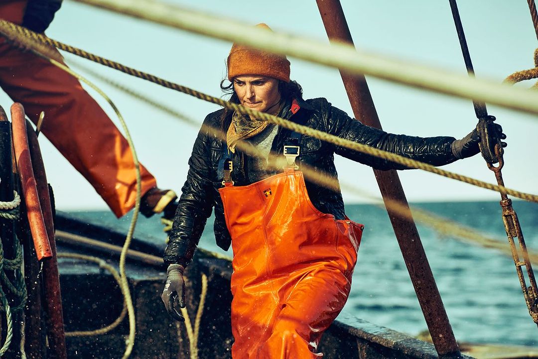 Woman in orange waders and a beanie on a weathered boat, holding ropes under a blue sky, possibly working as a fisherwoman or dockworker.
