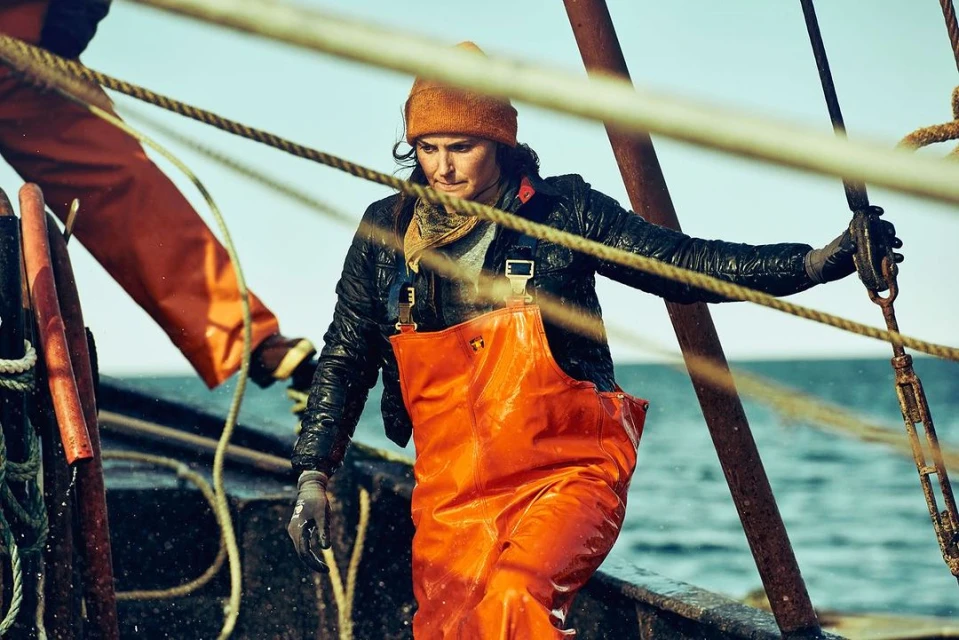 Woman in orange waders and a beanie on a weathered boat, holding ropes under a blue sky, possibly working as a fisherwoman or dockworker.