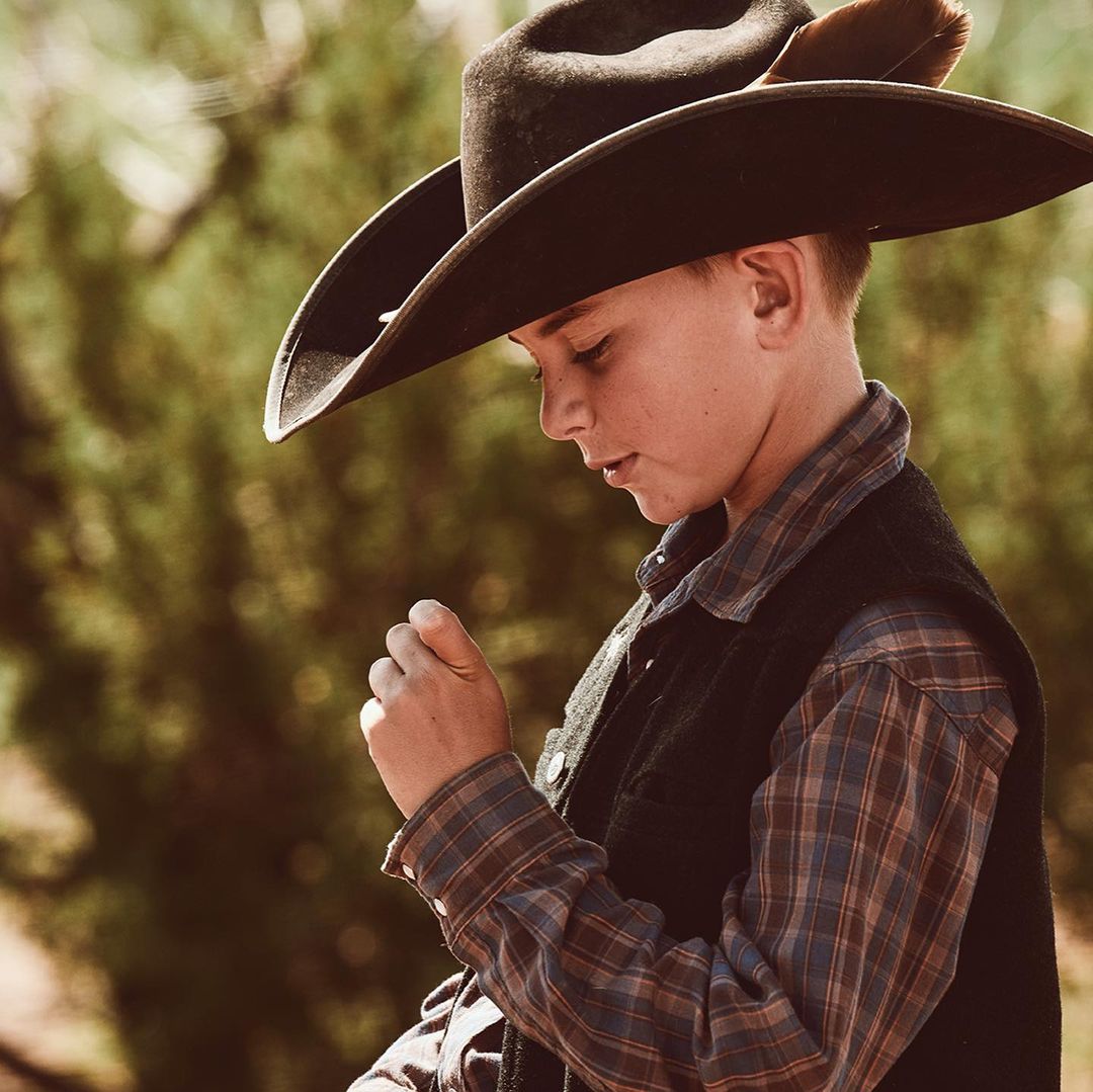 Young boy wearing cowboy hat, plaid shirt, and vest. Outdoors portrait with soft natural light.