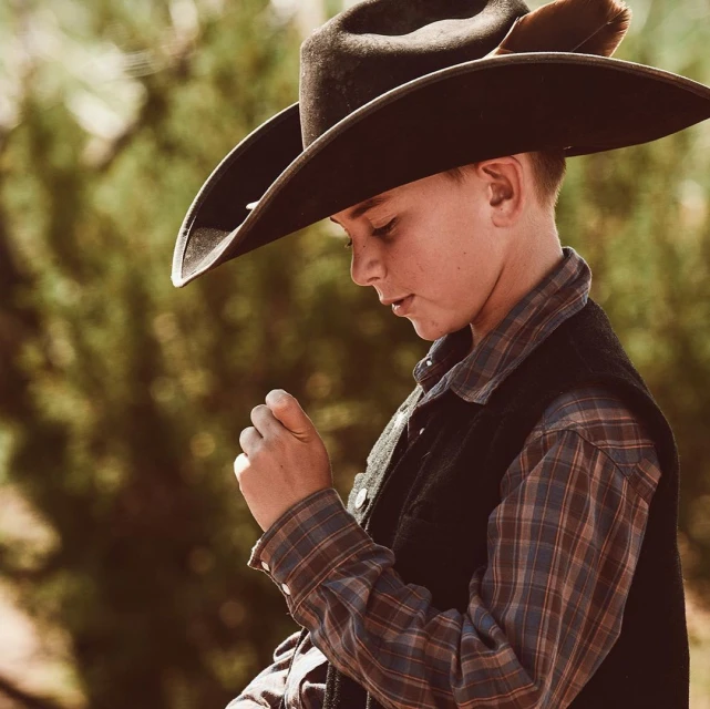 Young boy wearing cowboy hat, plaid shirt, and vest. Outdoors portrait with soft natural light.