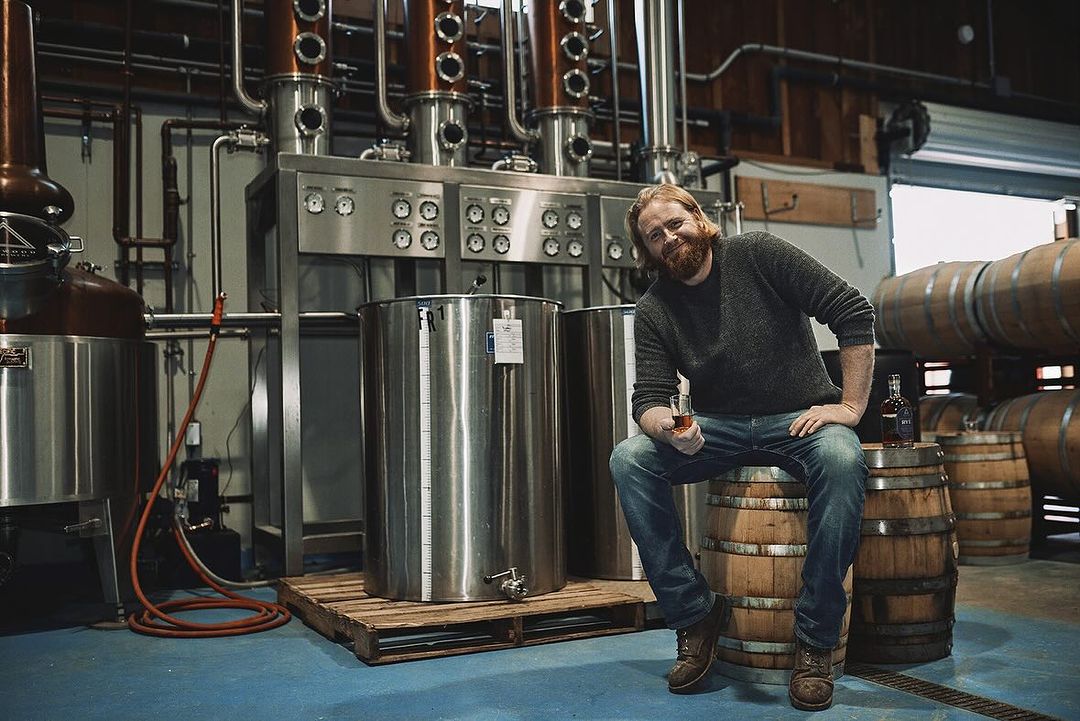Man with beard sits on barrel in distillery holding a glass of amber liquid. Distilling equipment and oak barrels visible.