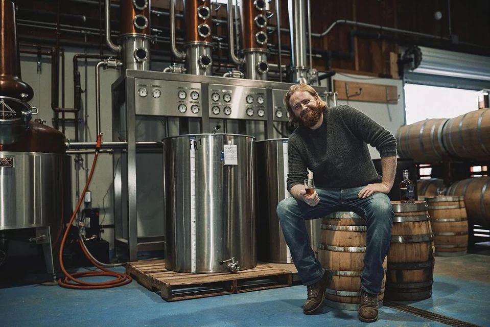 Man with beard sits on barrel in distillery holding a glass of amber liquid. Distilling equipment and oak barrels visible.