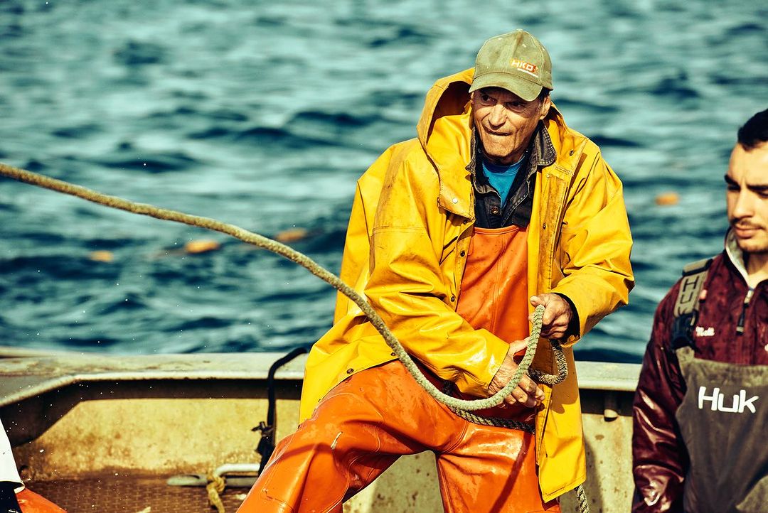A fisherman in a yellow raincoat pulling a rope on a boat with another man standing nearby in front of a blue water background.