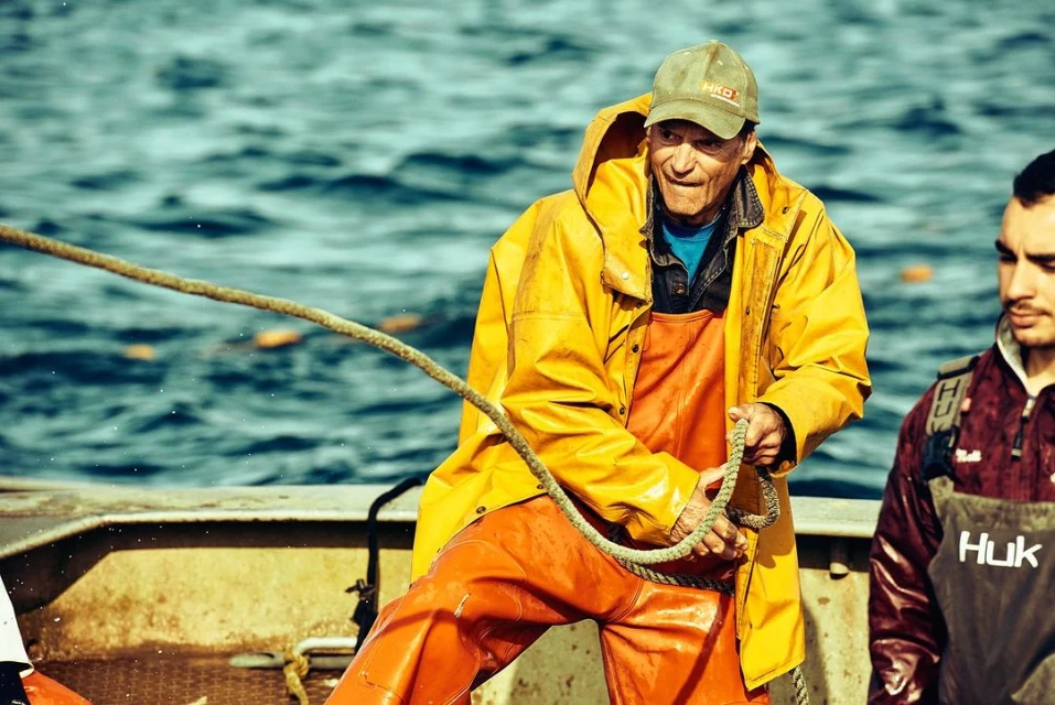 A fisherman in a yellow raincoat pulling a rope on a boat with another man standing nearby in front of a blue water background.