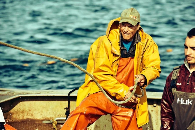 A fisherman in a yellow raincoat pulling a rope on a boat with another man standing nearby in front of a blue water background.