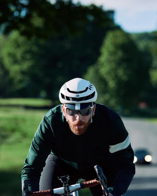 Adult cyclist with sunglasses and helmet rides a bicycle on a rural road with green trees and a car in the background on a sunny day.