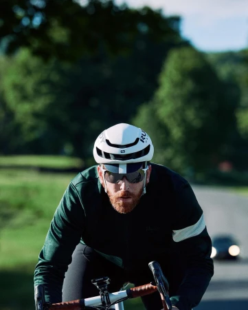Adult cyclist with sunglasses and helmet rides a bicycle on a rural road with green trees and a car in the background on a sunny day.