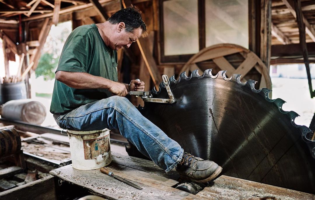 Adult male sharpening large saw blade indoors. Man in green shirt and jeans sits on bucket in rural workshop or barn.