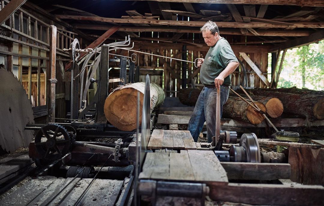 An older man works a vintage sawmill cutting a log inside of a rustic wooden building filled with equipment under natural light.