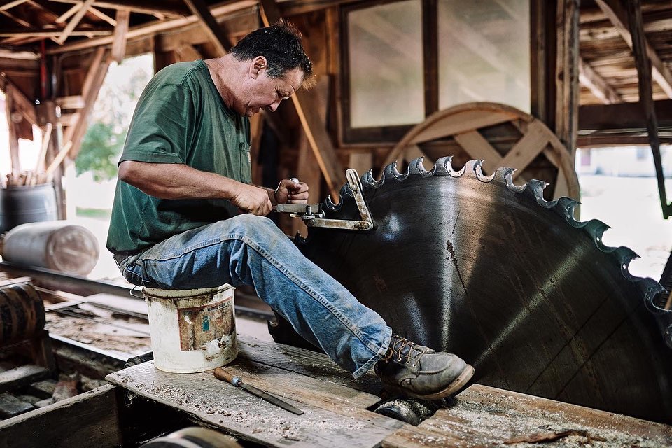 Adult man sharpens a massive saw blade indoors, sitting on a bucket with a file and vise against rustic wooden walls and sawdust.