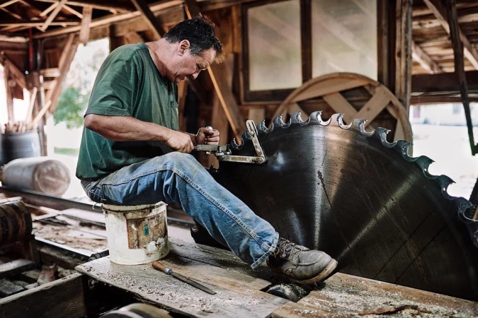 Adult man sharpens a massive saw blade indoors, sitting on a bucket with a file and vise against rustic wooden walls and sawdust.