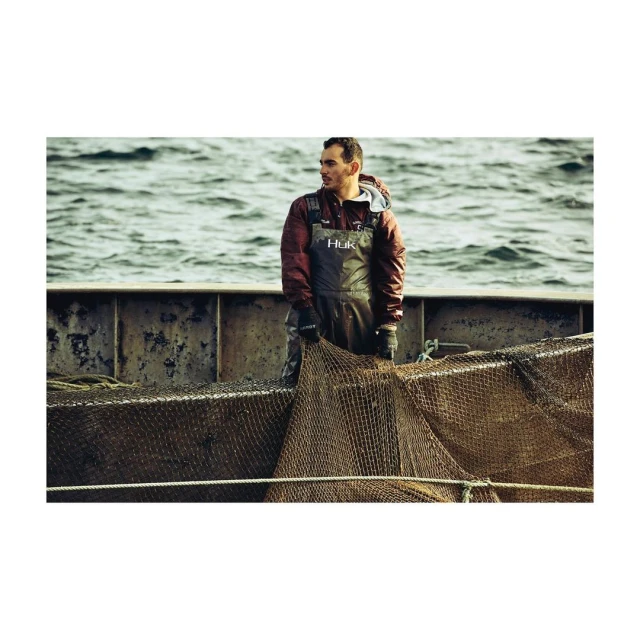 Man in fishing waders holding a fishing net on a boat. Ocean and waves in the background. Wearing dark gloves. Huk branded apparel.
