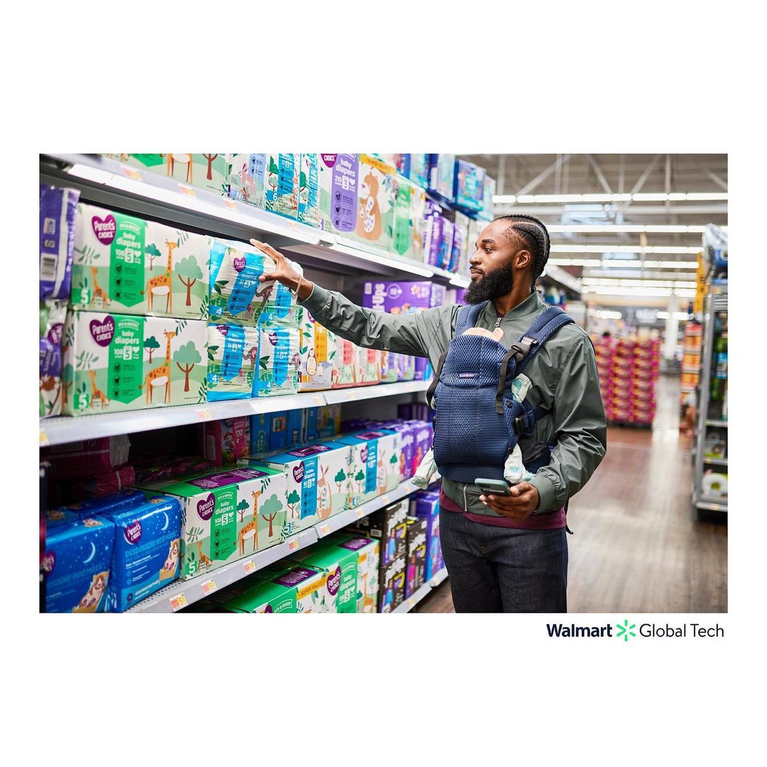 Adult male wearing a baby carrier in a grocery store aisle, reaching for a package of diapers on a shelf, holding smartphone.