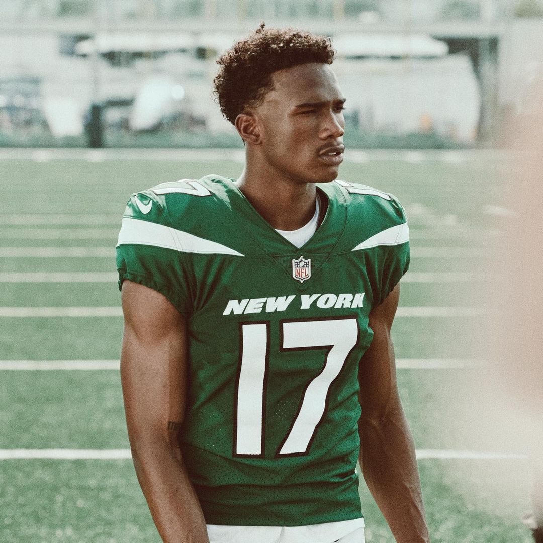 A male athlete in a New York Jets jersey stands on a green field, looking away with a serious expression. The image has a medium shot.