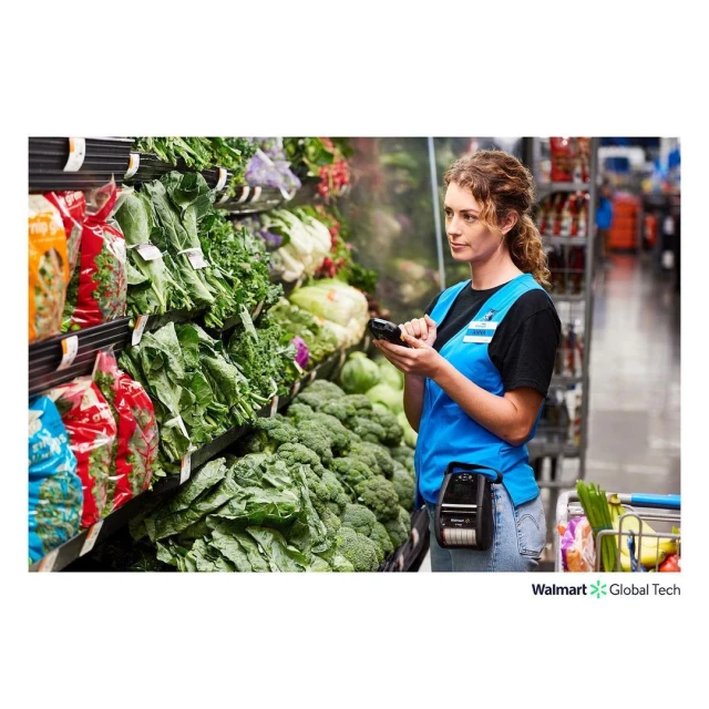 Adult woman in a blue vest scans produce in a grocery store, holding a scanner and wearing a belt-mounted device in an indoor setting.