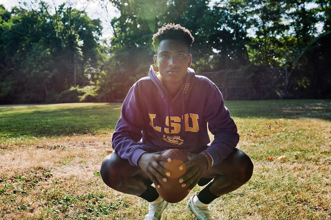 Portrait of a young adult male crouching in a grassy field, wearing an LSU hoodie, and holding a football in an outdoor setting.