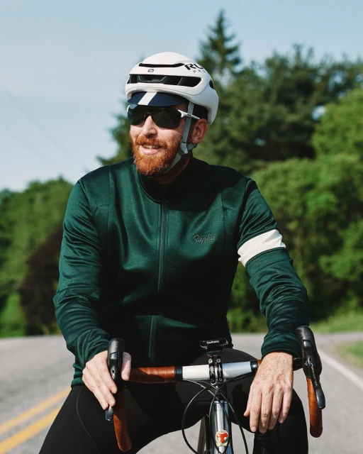 A cyclist pauses on a road, wearing a white helmet, green cycling jersey, and sunglasses, with trees and sky visible in the background.