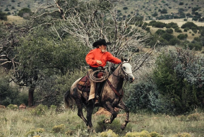 Cowboy on horseback gallops through a grassy, arid landscape with scattered trees under an overcast sky in a medium shot.