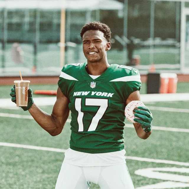 Football player in uniform poses with a coffee and donut on the field, smiling and looking off-camera in a medium shot.