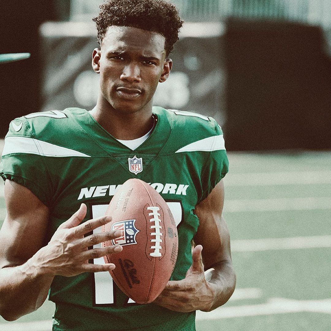 Close-up of an African-American football player in a green jersey holding a football, looking at the camera with a neutral expression.