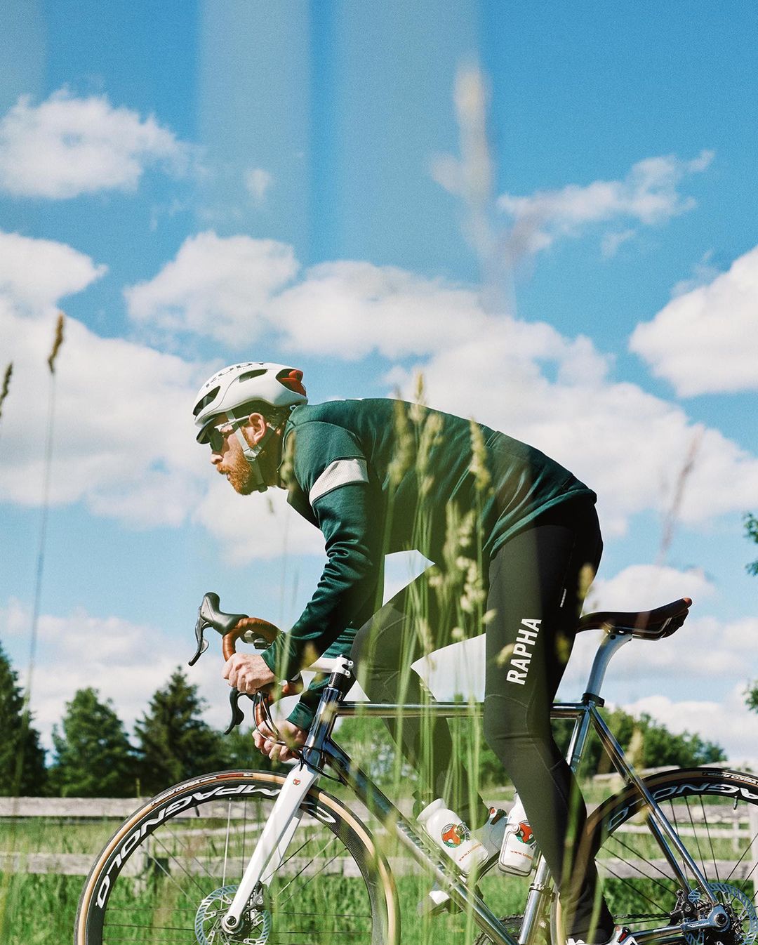 A cyclist rides a road bike on a rural road under a blue sky with clouds, wearing a helmet and athletic gear, captured on film.