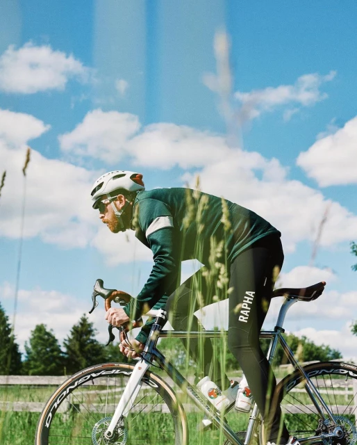 A cyclist rides a road bike on a rural road under a blue sky with clouds, wearing a helmet and athletic gear, captured on film.