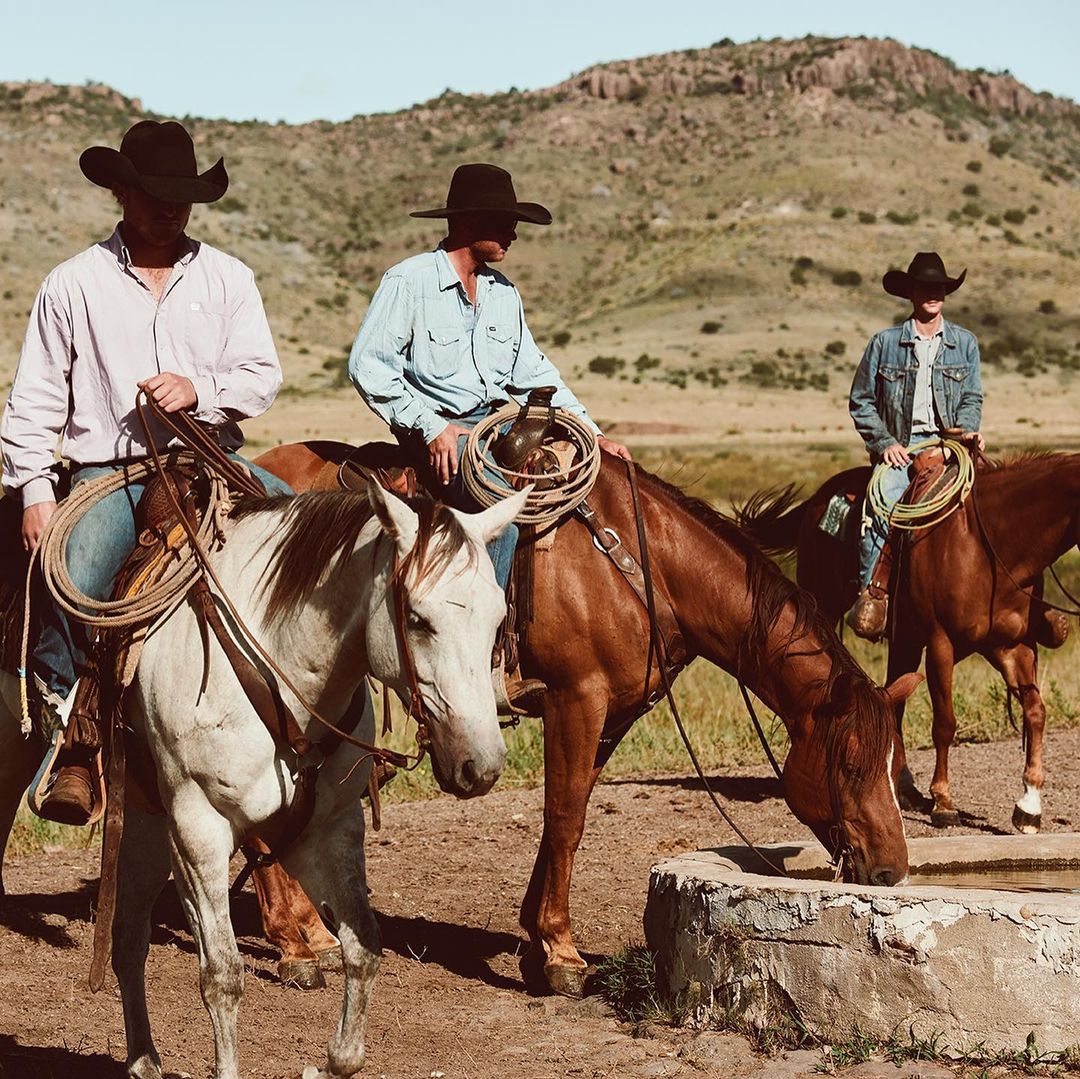 Three cowboys on horseback in a rural setting with rolling hills, wearing hats and casual clothing, creating a Western lifestyle image.