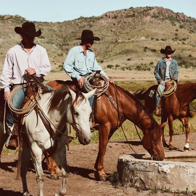 Three cowboys on horseback in a rural setting with rolling hills, wearing hats and casual clothing, creating a Western lifestyle image.