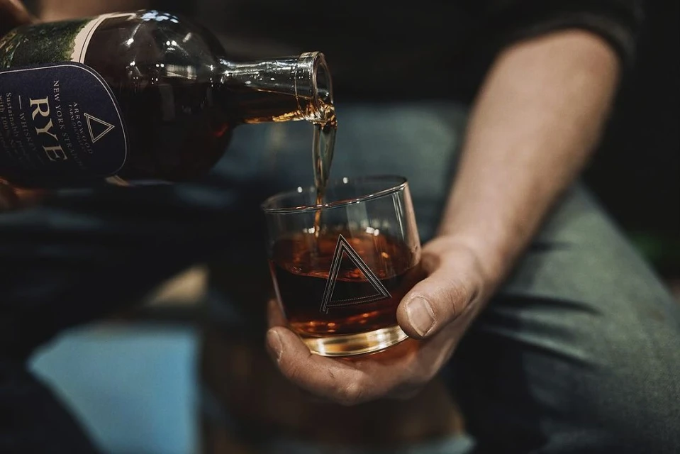 A person pours amber-colored rye whiskey from a bottle into a glass held in their other hand, with a shallow depth of field.