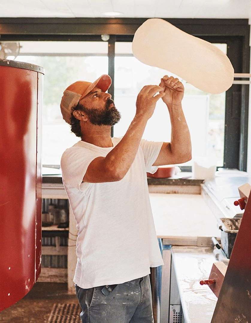 Adult male tossing pizza dough in a kitchen. He's wearing a t-shirt and hat, with a pizza oven visible. Natural light fills the space.