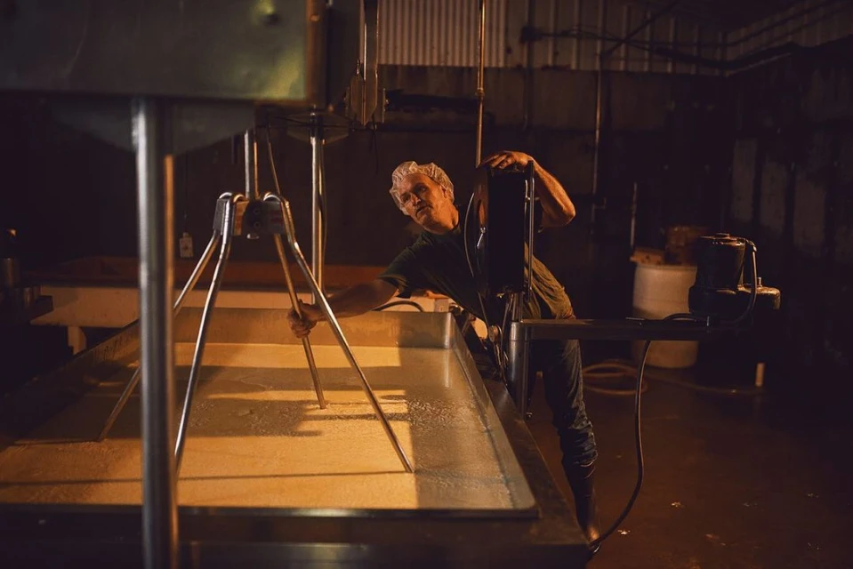 Man stirring a large vat of liquid with a metal tool in a dimly lit industrial setting, potentially processing food or ingredients.