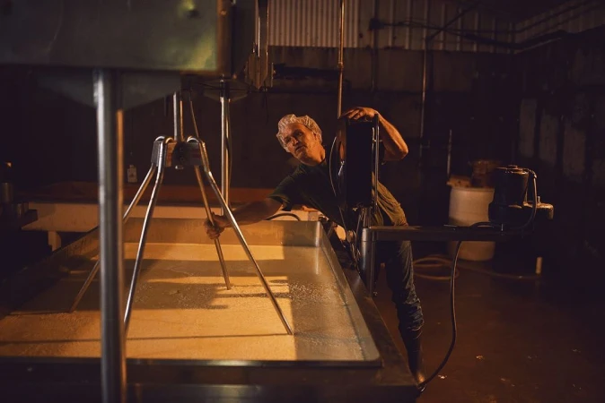 Man stirring a large vat of liquid with a metal tool in a dimly lit industrial setting, potentially processing food or ingredients.