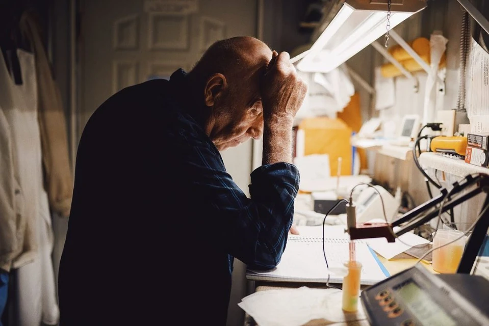 An elderly man sits indoors at a cluttered desk, his head in his hands, in a dimly lit, indoor setting with various tools and equipment.