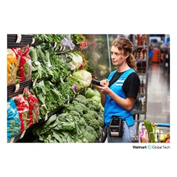 Retail worker in a blue vest holding a handheld scanner in the produce aisle by fresh vegetables