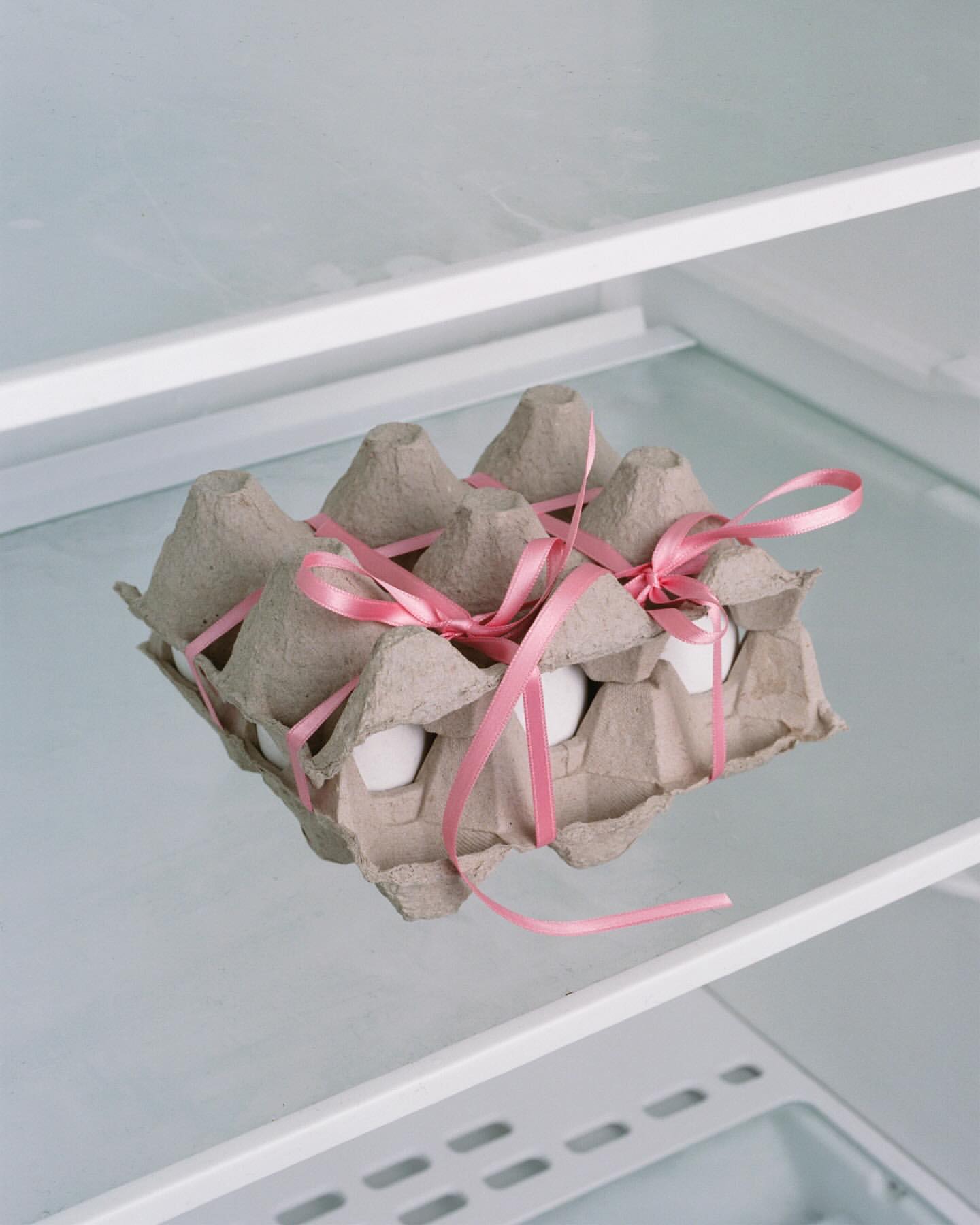 Egg carton with pink ribbon inside a refrigerator. The carton holds a dozen white eggs, displayed in a clean, bright refrigerator setting.